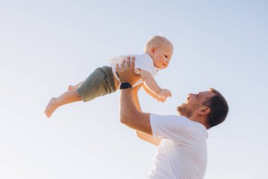 Neşeli bir baba bebeğini havaya kaldırır ve açık mavi gökyüzüne karşı yürek ısıtan bir an yaratır. Bu görüntü aile sevgisini ve mutluluğunu yansıtıyor..