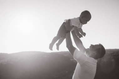 Güneşli bir arka planda bebeğini taşıyan bir babanın dokunaklı siyah beyaz görüntüsü..