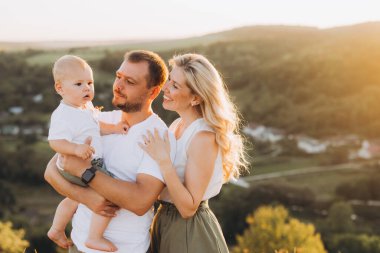 Üç kişilik neşeli bir aile, ebeveynleri ve bebekleri de dahil, güzel bir kırsal alanda huzurlu bir gün batımının tadını çıkarıyorlar, aşk ve beraberlik anları yakalıyorlar..
