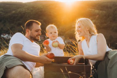 Üç kişilik neşeli bir aile, altın gün batımında elmaları ve kahkahaları paylaşarak pikniğin tadını çıkarıyor. Huzurlu manzara sıcak ve mutlu atmosfere ekleniyor..