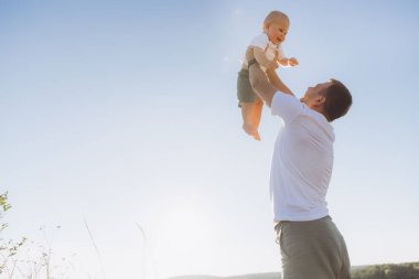 Bir baba gülümseyen bebeğini sevinçle havaya kaldırır, açık mavi gökyüzünün altında doğa ile çevrilidir. Babalığın ve kaygısız mutluluğun özünü yakalar..