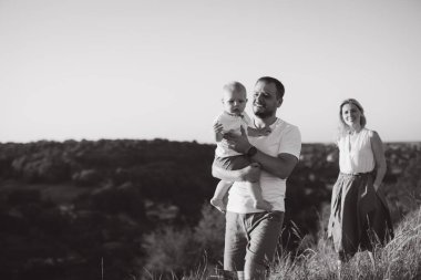 Doğada çekilen neşeli bir aile sahnesinde anne arka planda gülümserken bir baba çocuğunu kucağına alır. Aile sevgisi ve birlikteliği iletmek için ideal..