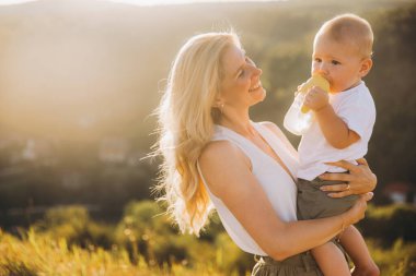 Bebeğini kucağında tutan bir annenin huzurlu bir sahnesi. Şişeden su içen, sıcak gün batımı ışığında yıkanan, sevgi ve şefkat taşıyan bir anne..