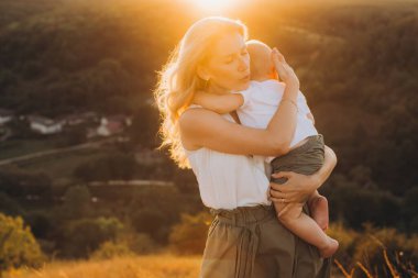 Bir anne gün batımında dışarıda dururken sevgiyle bebeğini kucağına alır. Sıcak parıltı, kırsal alanda huzurlu ve içten bir an yaratır..