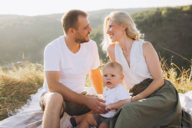 Güneşli bir günde, güzel bir doğal ortamda battaniyenin üzerinde oturup, aşkı, birlikteliği ve mutluluğu sembolize eden üç kişilik neşeli bir aile..