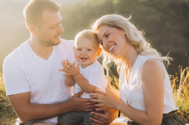 Neşeli bir aile ve gülümseyen bir bebek açık havada güneşli bir günün tadını çıkarıyor, doğada mutluluk ve beraberlik anları yakalıyor..