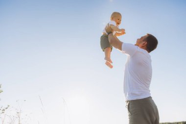 Bir baba mutlu bir şekilde gülümseyen bebeğini açık mavi gökyüzüne kaldırır, güneşli bir günde aşkı, oyunculuğu ve mutluluğu simgeler..