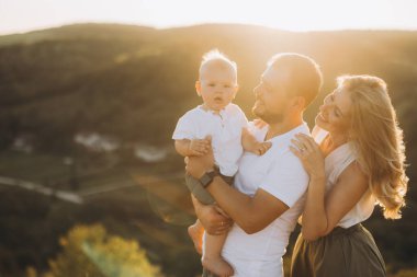 Neşeli bir aile ve bir erkek bebek güzel bir manzarayla çevrili açık havada güneşli bir günün tadını çıkarıyorlar. Ebeveynler çocuklarını kucaklıyor, neşeli ve sevgi dolu bir anı yakalıyorlar..