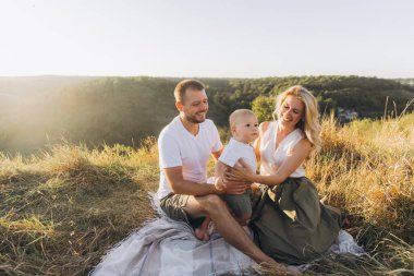 Neşeli bir aile, güneşli bir gün pikniğinde battaniyeye oturup bebekleriyle vakit geçirir. Manzara huzurlu ve güzel bir zemin sunar..