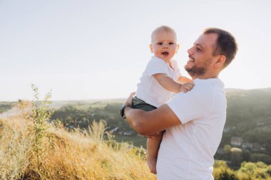 Neşeli bir baba doğada güneşli güzel bir günün tadını çıkarırken bebeğini kucağına alır. Bu barışçıl ortamda bağ ve mutluluk aşikar..