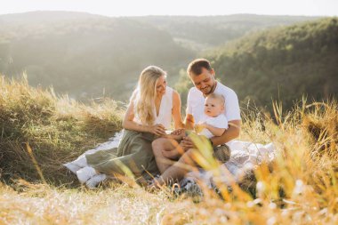 Neşeli bir aile dışarıda iyi vakit geçiriyor. Gülümseyen ebeveynler güneşli bir günde bebeğiyle birlikte piknik battaniyesinde oturur..