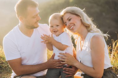 Neşeli bir aile açık havada güneşli bir günün tadını çıkarıyor. Gülümseyen ebeveynler ve mutlu bir bebek doğada sıcak güneş ışığıyla çevrili güzel bir anı paylaşırlar..