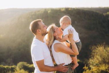 Neşeli bir aile açık havada güneşli bir günün tadını çıkarıyor. Ebeveynler çocuklarına sevgiyle sarılırlar güzel doğal bir manzara arkaplanı karşısında, mutluluk ve bağlantı anlarını yakalarlar..