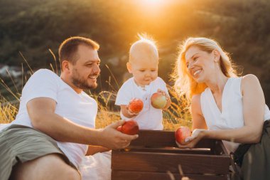 Üç kişilik neşeli bir aile güneşli bir pikniğin tadını çıkarıyor, dışarıda huzurlu bir ortamda elmalarını paylaşıyor ve gülümsüyor. Sıcak güneş ışığı neşeli atmosferi güçlendirir..