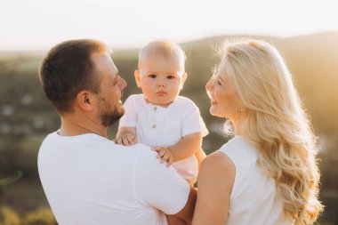 Neşeli bir aile gün batımında, anne babaların bebeklerini kucağında olduğu açık havada sevgi dolu bir anı paylaşır. Sıcak ışık, mutluluklarını ve bağlantılarını vurgular..