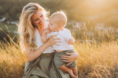 Güneşli bir alanda bebeğini kucağında tutan mutlu bir anne, altın saat boyunca ailesinin, sevginin ve doğanın sıcaklığını yakalıyor..