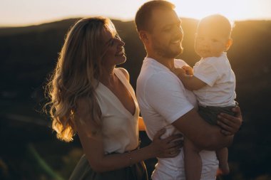 Üç kişilik neşeli bir aile dışarıda altın güneş ışığını emiyor. Ebeveynler güzel bir günbatımının keyfini çıkarırken bebeklerini kucaklarlar, sıcaklığı, sevgiyi ve birlikteliği aktarırlar..