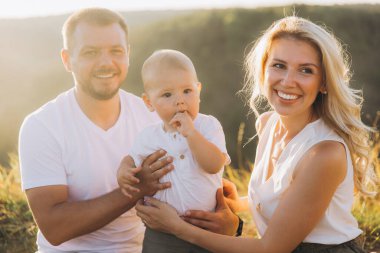 Neşeli bir aile gün ışığında dışarıda poz veriyor, gülümseyen ebeveynler ve sevimli bebekleri var. Sahne sıcaklık, neşe ve beraberlik saçıyor..