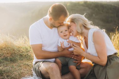 Mutlu bir aile sahnesi, anne babaların bebeklerini sevgiyle kucakladıkları, sıcak, güneşli bir açık hava ortamında yakalandıkları. İmaj mutluluk, sevgi ve beraberliği doğanın ortasında yayar..