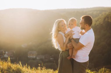 Neşeli bir aile sahnesi. Anne ve baba gülümseyen bebeklerini kucağında tutuyor. Sıcak güneş ışığında güzel doğal bir manzarayla çevrili..