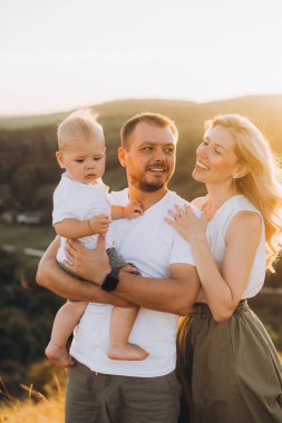 Neşeli bir aile açık havada güneşli bir günün tadını çıkarıyor. Ebeveynler, sıcak bir şekilde gülümseyerek, çocuklarını sevgi, mutluluk ve birlikteliği yansıtan, pitoresk bir manzaranın ortasında tutarlar..