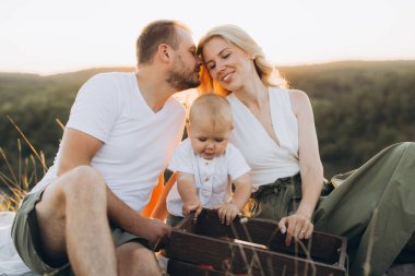 Neşeli bir aile sahnesinde ebeveynler ve yürümeye başlayan çocukları gün batımında, doğada sevgi, beraberlik ve sükuneti yakalıyorlar..