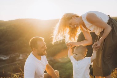 Üç kişilik neşeli bir aile güzel bir gün batımının tadını çıkarıyor. Ebeveynler çocuğun ellerini tutarken sıcak bir şekilde gülümserler, bir sevgi ve beraberlik anı yakalarlar..