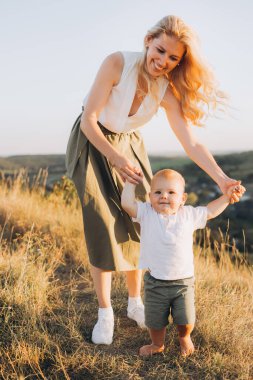 Neşeli bir anne bebeğinin çimenli bir arazide yürümesine yardım eder, güneşli bir günün tadını çıkarır. Sahne, doğada aile bağının güzel bir anını yakalar..