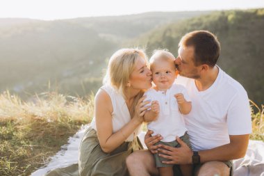 Sevgi dolu ebeveynler güneşli bir günde dışarıda dinlenirken çocuklarını kucaklayıp gülümserler. Aile sevgisini ve mutluluğu doğal bir ortamda yakalamak için mükemmel bir an..