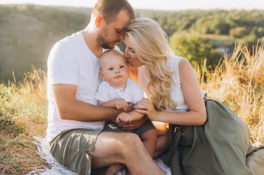 Güneşli bir çayırda oturan üç kişilik bir aileyle dokunaklı bir an. Sıcaklığı, sevgiyi ve güzel bir doğal ortamdaki bağlantıyı yakalıyorlar..