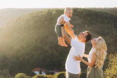 Doğanın tadını çıkaran neşeli bir aile. Anne gülümserken baba bebeği kaldırır. Güneşli bir günde dağlarda mutluluğu ve birlikteliği yakalamak..