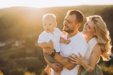 Güneşli bir günde ebeveynlerin bebeklerini kucağında tuttukları neşeli bir aile anı. Resim mutluluğu, aşkı ve birlikteliği tasvir ediyor..