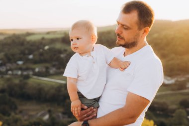 Bir baba gün batımında bebeğini sevgiyle kucağına alır ve nefes kesici kırsal manzaranın ortasında bağlarını gösterir..