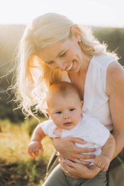 Gülümseyen bir anne sevgiyle bebeğini açık havada güneşli bir ortamda tutar. Kırsal kesimde neşeli bir bağ ve sıcaklık anı yakalar..