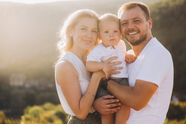 Neşeli bir aile açık havada güneşli bir günün tadını çıkarıyor. Gülümseyen anne ve babalar sevimli bebeklerini kucağında tutarak samimiyet ve beraberlik yayıyorlar..