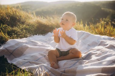 Güneşli bir tarlada piknik battaniyesinde oturan sevimli bebek, sıcak bir günde doğanın güzelliğini merak ve neşe ile yaşıyor..