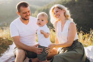 Küçük oğluyla gülümseyen ebeveynler açık havada güneşli bir günün tadını çıkarıyorlar. Aile doğal bir ortamda battaniyeye oturmuş sıcak ve neşeli bir anı yakalıyor..