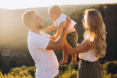 Neşeli aile gün batımında dışarıda kaynaşıyor. Anne ve baba bebeklerini tutarken gülümsüyor, birlikte güzel doğal bir ortamda değerli anların tadını çıkarıyorlar..