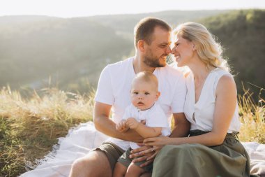 Güneşli bir günde tepede oturan bebekli gülümseyen ebeveynler. Aile sevgisini yakalamak ve doğanın güzelliği arasında bağ kurmak.