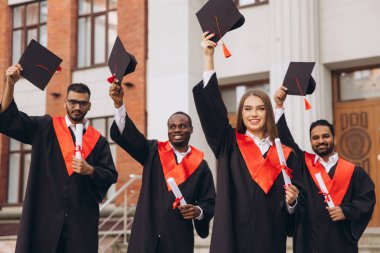 Bir üniversite binasının önünde çeşitli mezunlar kutlama yapıyor, diplomaları tutuyor ve havada şapkalar yükseltiyor, başarı ve neşe ifade ediyorlar..