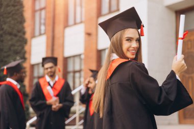 Mutlu genç bir kadın arkadaşlarıyla birlikte mezun oluyor, akademik elbiseler ve havan mermileriyle dışarıda kutlama yapıyor, özel bir günde neşe ve başarıyı ifade ediyor..