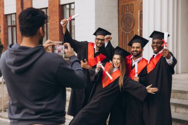 Bir üniversite binası önünde diplomalarıyla poz veren çeşitli mezunlardan oluşan neşeli bir grup, başarılarının ve akademik başarılarının anılarını yakalıyorlar..
