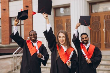 Üç farklı mezun dışarıda başarılarını kutluyor, ellerinde şapkalar ve diplomalar var. Onların ifadeleri mezuniyet gününde neşe ve başarıyı yansıtır..