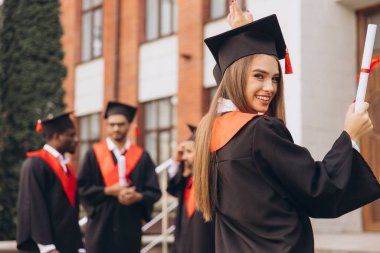 Mutlu genç bir kadın arkadaşlarıyla birlikte mezun oluyor, akademik elbiseler ve havan mermileriyle dışarıda kutlama yapıyor, özel bir günde neşe ve başarıyı ifade ediyor..