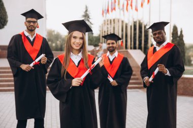 Mezuniyetlerini sevinçle kutlayan şapkalı ve cüppeli bir grup öğrenci ellerinde diplomalarla. Arka planda bayraklarla açık havada yakalanmış..