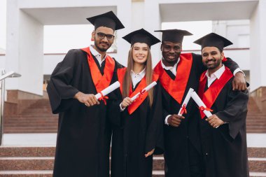 Çeşitli mezunlardan oluşan neşeli bir grup kep ve cübbe içinde akademik başarılarını dışarıda kutluyor, diplomalarını tutuyor ve kameraya gülümsüyor..