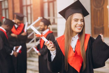 Mutlu genç bir kadın arkadaşlarıyla birlikte mezun oluyor, akademik elbiseler ve havan mermileriyle dışarıda kutlama yapıyor, özel bir günde neşe ve başarıyı ifade ediyor..