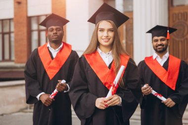 Bir grup genç mezun, başlıkları ve cübbeleriyle güvenle gülümsüyor, bir üniversite kampüsünün dışında diplomalarını tutuyor, akademik başarılarını kutluyorlar..