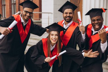 Çeşitli mezunlardan oluşan neşeli bir grup başarılarını kutluyor ve ellerinde diplomalar var. Mezuniyet gününde açık havada başarı ve başarının neşeli ifadesi..