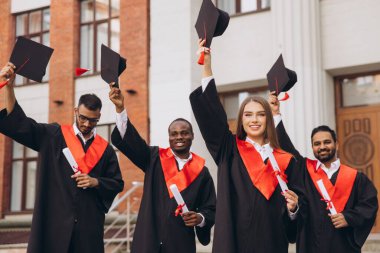 Bir üniversite binasının önünde çeşitli mezunlar kutlama yapıyor, diplomaları tutuyor ve havada şapkalar yükseltiyor, başarı ve neşe ifade ediyorlar..